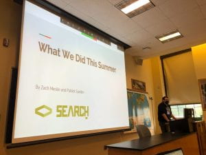 In a classroom a projector screen displays the words "What we did this summer" with a green logo that says SEARCH. A man in a black hoodie stands to the right near a computer.