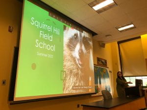 In a classroom a projector screen displays the words "Squirrel Hill Field School" with a picture of a raccoon holding a trowel. A woman in a dress stands to the right near a computer.