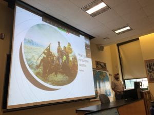 In a classroom a projector screen displays the words "What I Did on my Summer Vacation" with a picture of him as Washington in the painting Washington crosses the Delaware. A man in a checkered shirt and ball cap stands to the right near a computer.