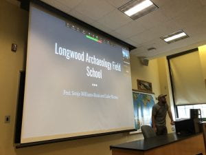 In a classroom a projector screen displays the words "Longwood Archaeology Field School." A man in a grey t-shirt and ball cap stands to the right near a computer.